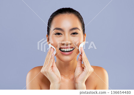 A woman is using cotton pads to clean her face. She is smiling and looking towards the camera. The setting has a plain light background. This scene shows a routine skincare activity. A woman is using cotton pads to clean her face. She is smiling and looking towards the camera. The setting has a plain light background. This scene shows a routine skincare activity. 136654480