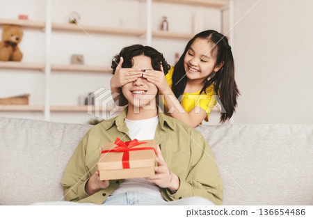 Smiling teen girl closes eyes of young asian man and present box with gift, sit on sofa in living room interior. Celebrate birthday, family relationships, fathers day, holiday due covid-19 pandemic Smiling teen girl closes eyes of young asian man and present box with gift, sit on sofa in living room interior. Celebrate birthday, family relationships, fathers day, holiday due covid-19 pandemic 136654486