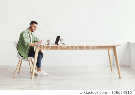 A young man sits at a wooden desk in a bright room. He wears headphones and writes in a notebook. A laptop and a cup are on the desk. The space feels open and inviting. 136654510