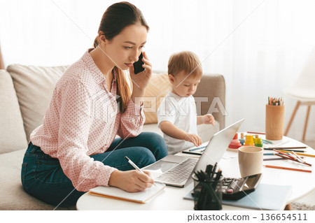 Work at maternity leave. Woman working at home and spending time with son, freelancer lady multitasking talking on cellphone and taking notes, free space Work at maternity leave. Woman working at home and spending time with son, freelancer lady multitasking talking on cellphone and taking notes, free space 136654511