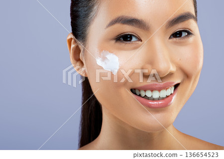 A woman smiles as she applies skincare cream to her face. She is indoors against a plain background. This activity highlights her skincare routine and personal care habits. A woman smiles as she applies skincare cream to her face. She is indoors against a plain background. This activity highlights her skincare routine and personal care habits. 136654523