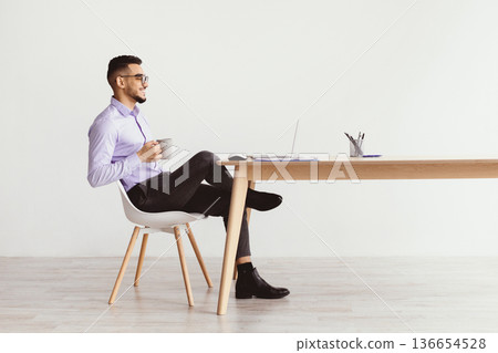 A man sits on a chair with a laptop open on a desk. He holds a large cup while looking at the screen. The room has bright walls and a minimalistic design, with stationery on the desk. A man sits on a chair with a laptop open on a desk. He holds a large cup while looking at the screen. The room has bright walls and a minimalistic design, with stationery on the desk. 136654528