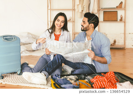 Travel concept. Travelers couple choosing touristic destination for vacation, holding map while sitting near open suitcase in living room. Young lovers planning next trip together Travel concept. Travelers couple choosing touristic destination for vacation, holding map while sitting near open suitcase in living room. Young lovers planning next trip together 136654529