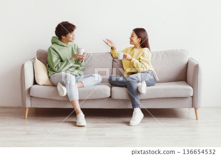 Young Asian boyfriend and girlfriend drinking coffee and chatting while sitting on sofa at home. Affectionate millennial couple enjoying hot drink, having conversation, spending time together 136654532