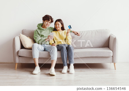 Cheery young Asian couple with credit card and smartphone sitting on sofa, ordering goods on internet, buying things in web store at home, copy space. Online shopping, distance banking concept Cheery young Asian couple with credit card and smartphone sitting on sofa, ordering goods on internet, buying things in web store at home, copy space. Online shopping, distance banking concept 136654546