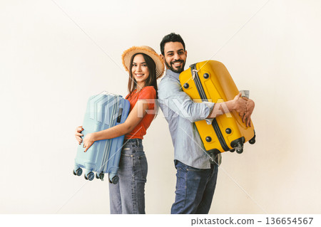 Excited young tourists couple ready for traveling abroad, holding suitcases in hands and smiling at camera, standing over light wall. Air flight journey concept 136654567