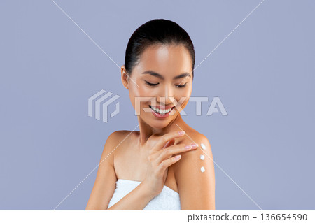 A woman stands in a studio with a plain background. She smiles as she applies cream to her shoulder. Her hair is tied back, and she is dressed in a towel. She seems focused on her skincare routine. 136654590