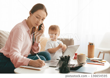 Young mother working at home while her son drawing next to her, freelancer lady talking on smartphone and taking notes, family spending time together at home, free space Young mother working at home while her son drawing next to her, freelancer lady talking on smartphone and taking notes, family spending time together at home, free space 136654595