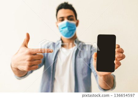Young arab man in protective medical mask pointing at smartphone with blank screen, guy demonstrating mockup for your add, app or website design, selective focus Young arab man in protective medical mask pointing at smartphone with blank screen, guy demonstrating mockup for your add, app or website design, selective focus 136654597