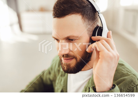 Bearded Man Wearing Wireless Headphones Listening To Music Online Looking Aside At Home. Headshot Of Guy Listens To Podcast Or Enjoying Favorite Song On Weekend. Side View 136654599