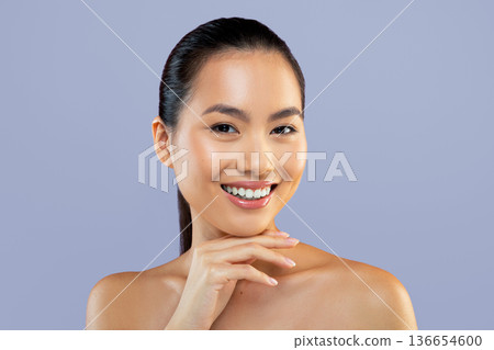 A woman with straight dark hair is smiling and resting her chin on her hand. She has clear skin and is positioned in front of a plain background. The scene shows positive energy. 136654600