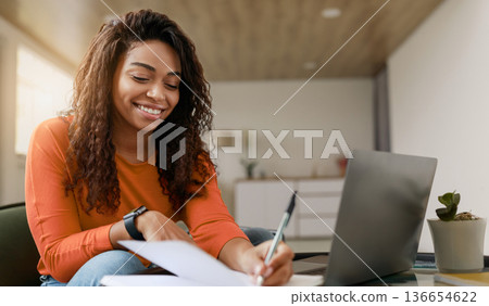 A woman writes notes on a piece of paper while seated at a table. She looks happy as she focuses on her work. A laptop is open in front of her. The room is bright and has a modern design. 136654622
