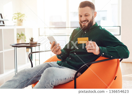 Mobile Shopping. Cheerful Man Using Phone And Credit Card Purchasing Things In Application Sitting In Chair At Home. Side View Of Guy Holding Bank Card And Smartphone Using Bank App Online Mobile Shopping. Cheerful Man Using Phone And Credit Card Purchasing Things In Application Sitting In Chair At Home. Side View Of Guy Holding Bank Card And Smartphone Using Bank App Online 136654623