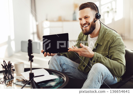 Happy Man Showing Digital Tablet Empty Screen To Smartphone Making Video Call Or Filming For Blog Sitting In Chair At Home. Modern Online Communication And Gadgets. Selective Focus Happy Man Showing Digital Tablet Empty Screen To Smartphone Making Video Call Or Filming For Blog Sitting In Chair At Home. Modern Online Communication And Gadgets. Selective Focus 136654641