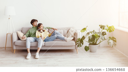 Positive young Asian couple relaxing on sofa, cuddling and looking out window in living room, free space. Millennial husband and wife chilling on comfy couch, spending time together 136654668