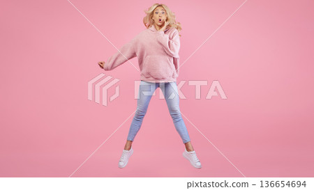 A girl wearing a pink sweatshirt and blue jeans jumps in a studio. She shows a surprised expression, with her hands near her face. The background is bright pink, enhancing the lively mood. A girl wearing a pink sweatshirt and blue jeans jumps in a studio. She shows a surprised expression, with her hands near her face. The background is bright pink, enhancing the lively mood. 136654694