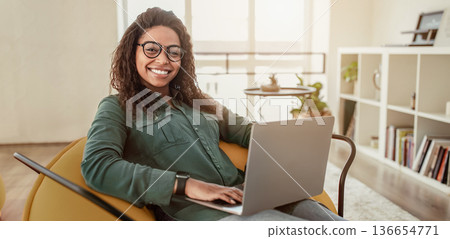 A young woman sits comfortably on a chair in a modern room. She works on her laptop and smiles while looking at the screen. The room has bright light and simple furniture. A young woman sits comfortably on a chair in a modern room. She works on her laptop and smiles while looking at the screen. The room has bright light and simple furniture. 136654771
