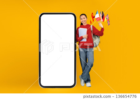A young woman stands next to a large empty smartphone. She holds flags from different countries and has papers in her hands. She smiles and wears casual clothes. The background is bright yellow. 136654776