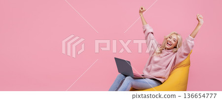 A young woman is sitting on a yellow bean bag chair in a room with a pink wall. She has long hair and is smiling with her arms raised in excitement. A young woman is sitting on a yellow bean bag chair in a room with a pink wall. She has long hair and is smiling with her arms raised in excitement. 136654777