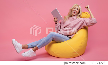 A woman sits in a yellow bean bag chair and smiles while holding a tablet. She looks happy and relaxed as she connects with something on the screen. Bright colors enhance the mood. 136654778