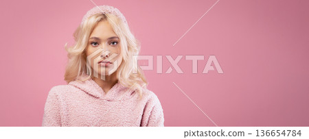 A young woman with blonde hair poses in front of a pink background. She wears a warm pink sweater. The focus is on her expression and the simple color setting to highlight her presence. 136654784
