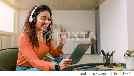 A woman is sitting on a chair in a modern living room. She is wearing headphones and smiling while having a video call on her tablet. The room is well lit with natural light. 136654785
