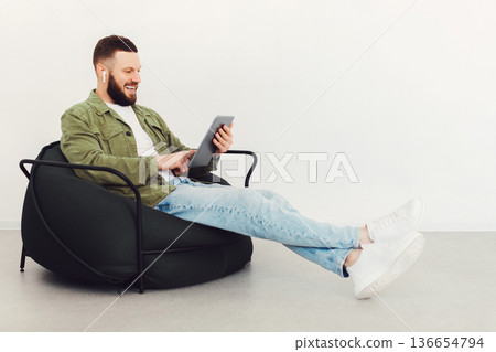 Side View Of Man Using Tablet Computer And Earbuds Earphones Watching Movie Online Sitting In Chair Over White Wall Indoors On Weekend. E-Learning, Internet And Technology Concept 136654794