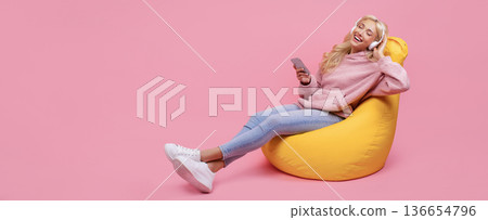 A woman with long hair is sitting on a yellow bean bag. She is smiling and listening to music with headphones. She holds a smartphone in one hand and relaxes in a pink room. 136654796