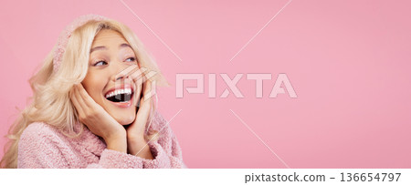 Woman with long light hair smiles widely while resting her hands on her cheeks. She is wearing a pink sweater and stands in front of a solid pink background. The setting is bright and cheerful. Woman with long light hair smiles widely while resting her hands on her cheeks. She is wearing a pink sweater and stands in front of a solid pink background. The setting is bright and cheerful. 136654797