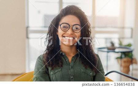 A woman with curly hair and glasses sits in a well-lit indoor area. She looks directly at the viewer with a smile, showing confidence and warmth. The space has a modern feel. A woman with curly hair and glasses sits in a well-lit indoor area. She looks directly at the viewer with a smile, showing confidence and warmth. The space has a modern feel. 136654838