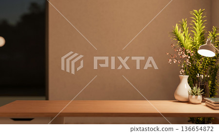 Books and potted plant and lamp on wooden table across the wall and glass window in dim living room. 136654872