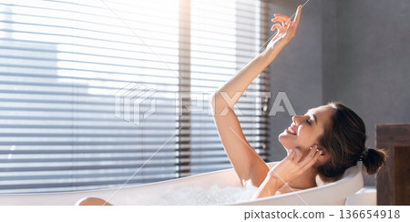 A woman sits in a bathtub filled with bubbles, enjoying the warm afternoon light. She raises her hand playfully while smiling. Sunlight streams through the blinds into the room. A woman sits in a bathtub filled with bubbles, enjoying the warm afternoon light. She raises her hand playfully while smiling. Sunlight streams through the blinds into the room. 136654918