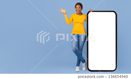 A woman stands next to a large blank screen. She is smiling and waving. The background is blue. She wears a yellow top, jeans, and white shoes. This scene suggests excitement and engagement. A woman stands next to a large blank screen. She is smiling and waving. The background is blue. She wears a yellow top, jeans, and white shoes. This scene suggests excitement and engagement. 136654940