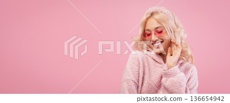 A young woman enjoys a moment while wearing a fluffy pink hoodie and pink sunglasses. She poses playfully in front of a pink background and has long blonde hair. A young woman enjoys a moment while wearing a fluffy pink hoodie and pink sunglasses. She poses playfully in front of a pink background and has long blonde hair. 136654942
