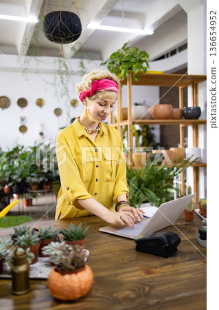 A smiling woman works on a laptop at a wooden counter in a plant shop, surrounded by greenery 136654952