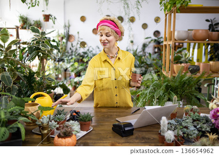 A woman in a yellow shirt holds a potted succulent in a plant shop, surrounded by greenery and other plants A woman in a yellow shirt holds a potted succulent in a plant shop, surrounded by greenery and other plants 136654962