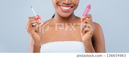 A woman stands smiling while holding a lip gloss in one hand and a lip applicator in the other hand. She appears happy and is in a bright setting, suitable for a beauty promotion. A woman stands smiling while holding a lip gloss in one hand and a lip applicator in the other hand. She appears happy and is in a bright setting, suitable for a beauty promotion. 136654963