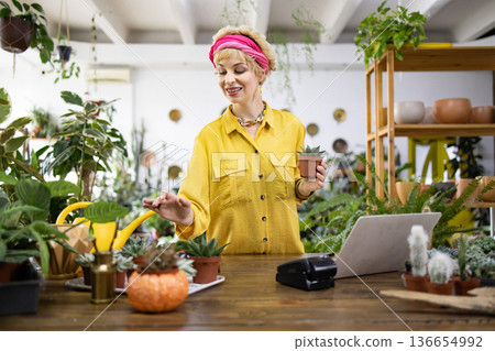 A woman in a yellow shirt holds a small succulent in a plant shop, surrounded by greenery A woman in a yellow shirt holds a small succulent in a plant shop, surrounded by greenery 136654992