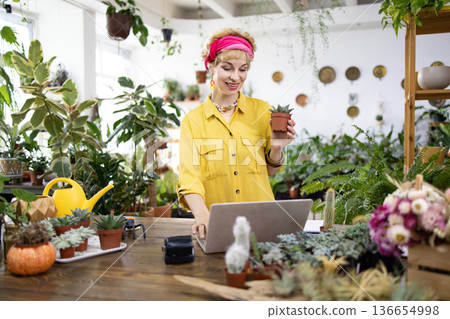 A woman in a yellow shirt holds a succulent while working on a laptop in a bright, inviting plant shop A woman in a yellow shirt holds a succulent while working on a laptop in a bright, inviting plant shop 136654998