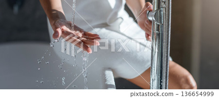 Person sits beside bathtub with water running from shower. The setting is a bathroom with natural light and modern fixtures. The scene captures a moment of personal care and relaxation. Person sits beside bathtub with water running from shower. The setting is a bathroom with natural light and modern fixtures. The scene captures a moment of personal care and relaxation. 136654999