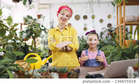 A woman and a young girl smile in a plant shop surrounded by greenery and a laptop on the table A woman and a young girl smile in a plant shop surrounded by greenery and a laptop on the table 136655019