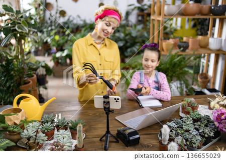 A mother and daughter are filming a gardening tutorial in a plant shop, surrounded by greenery and tools 136655029