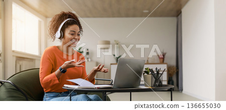 A woman sits on a chair with headphones on, smiling as she speaks on a laptop. There are plants and stationery on a table nearby, providing a bright setting for her conversation. 136655030