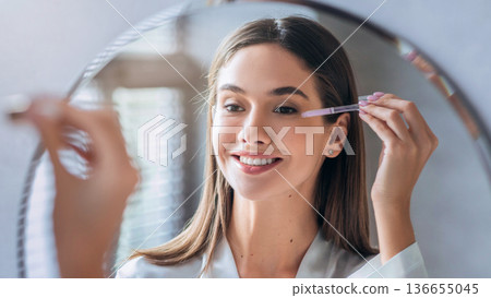 A woman smiles as she applies makeup in front of a mirror. She uses a makeup tool to define her eyes. Natural light fills the room, highlighting her focused expression. A woman smiles as she applies makeup in front of a mirror. She uses a makeup tool to define her eyes. Natural light fills the room, highlighting her focused expression. 136655045