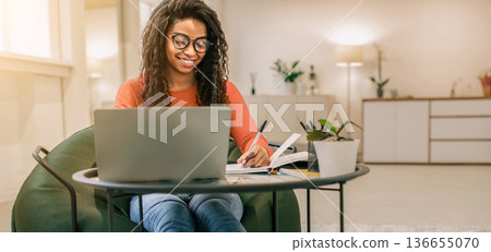 A young woman sits at a round table in a well-lit room. She smiles while writing notes and looking at a laptop. A small plant sits nearby, adding to the cozy atmosphere of the space. A young woman sits at a round table in a well-lit room. She smiles while writing notes and looking at a laptop. A small plant sits nearby, adding to the cozy atmosphere of the space. 136655070
