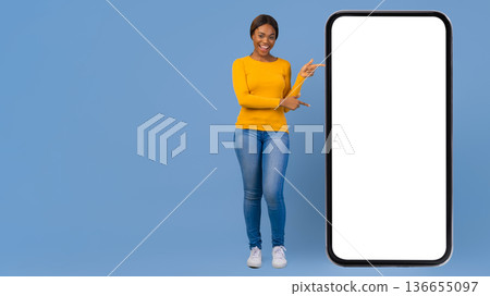 A person stands in front of a blue wall holding a blank smartphone. She smiles and points to the screen. The setting is bright and inviting, showing technology use in daily life. 136655097