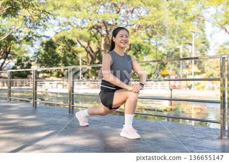 Pretty asian woman in sportswear crouching stretching legs muscles aside railing on pavement in park 136655147