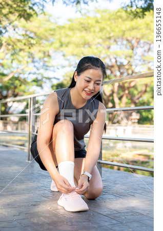 Pretty asian woman in sportswear sitting crouching tying sneaker's lace on concrete pavement in park Pretty asian woman in sportswear sitting crouching tying sneaker's lace on concrete pavement in park 136655148