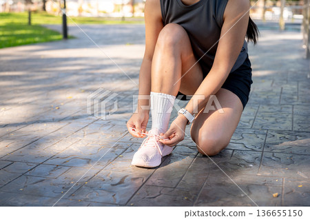 Close up of woman in sportswear sitting crouching tying sneaker's lace on concrete pavement in park. Close up of woman in sportswear sitting crouching tying sneaker's lace on concrete pavement in park. 136655150