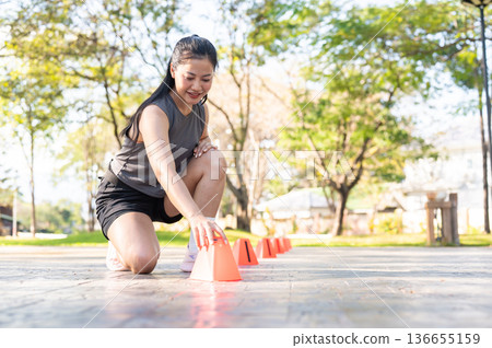 Pretty asian woman in sportswear setting up cones before exercise on pavement in park under sunlight Pretty asian woman in sportswear setting up cones before exercise on pavement in park under sunlight 136655159